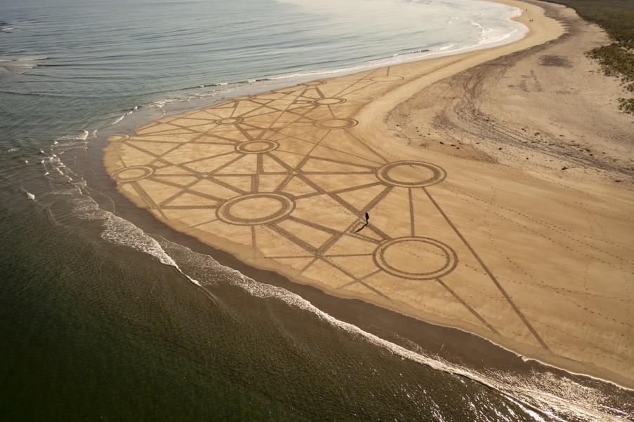 A man makes a geometric design on a sandy beach as the tide comes in.