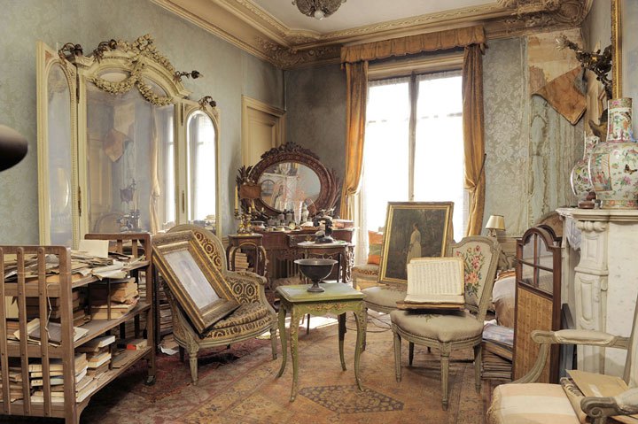 Interior of a high-end apartment, filled with art nouveau style furnishings including a mirrored vanity and several chairs. The furnishings are high-end, but are covered in dust. 