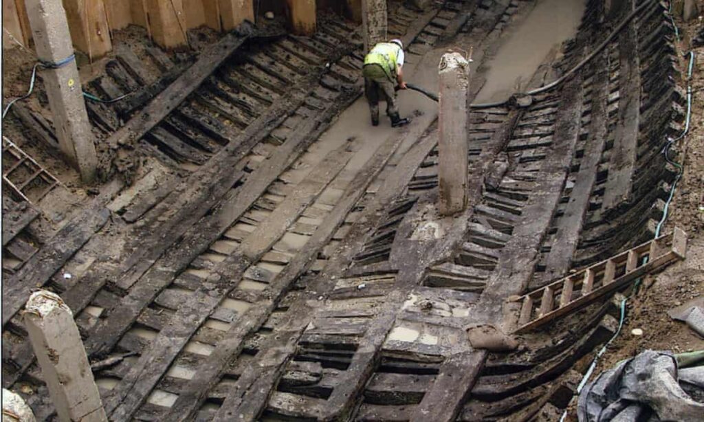 An archeaological excavation showing the interior structure of a large wooden ship. An archeaologist removes silt from the bottom of the ship with a large vacuum. 