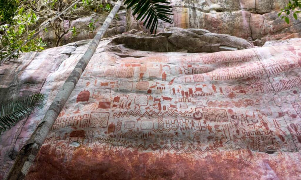 A cliff face features numerous rock art images in red ochre. The drawings are tightly packed together and extend many meters upwards. 