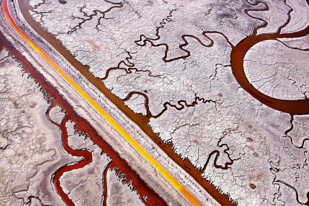 An aerial photograph of a wetland. The water is a rusty red color and it makes pathways through the whitish grey soil. 