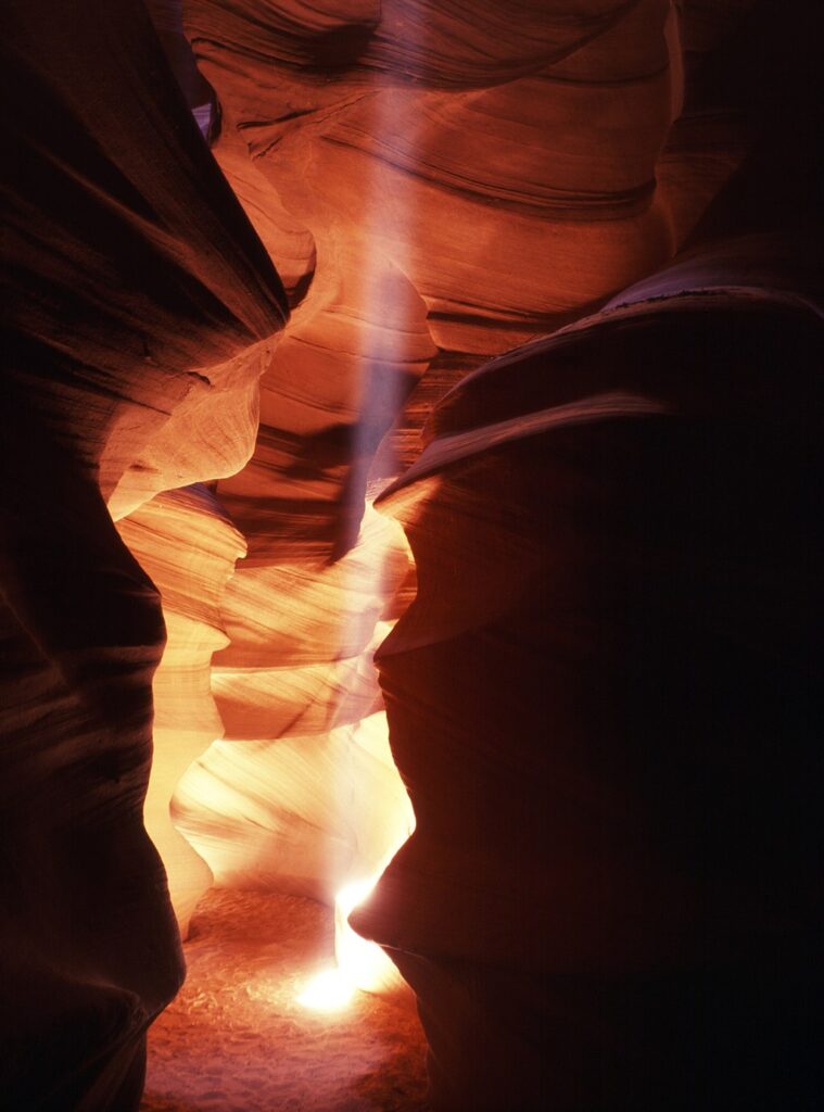 A shaft of light breaks through a desert canyon. The walls of the canyon have been carved by water and are various shades of yellow, orange, and brown. 