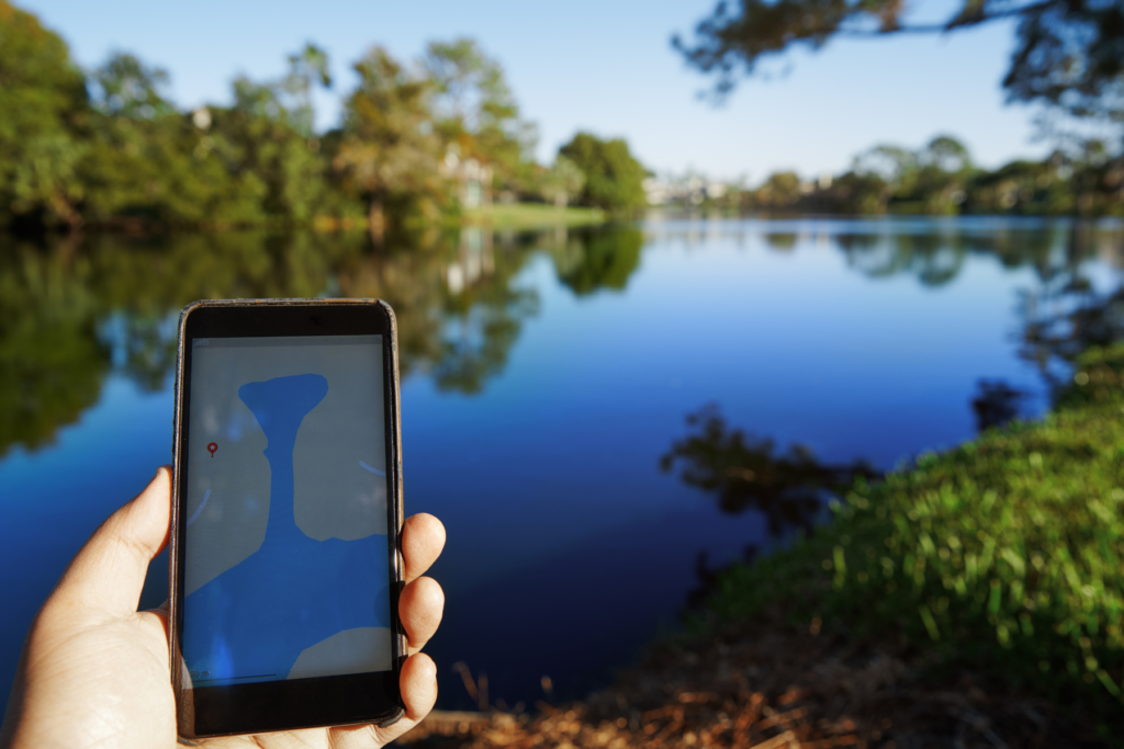 A person's hand holds a phone. In the background there's a lake surrounded by a forest.