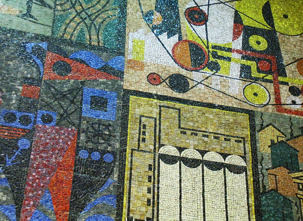 A close up of an abstract mural down in glass mosaic. The mural features geometric shapes in black, blue, red, yellow and beige. 