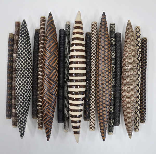 A set of cylinderical ceramic forms, arranged in a row. Each of the cylinders is carved with different geometric patterns and glazed in brown, white, and black. Some of the cylinders are brought to a point. 