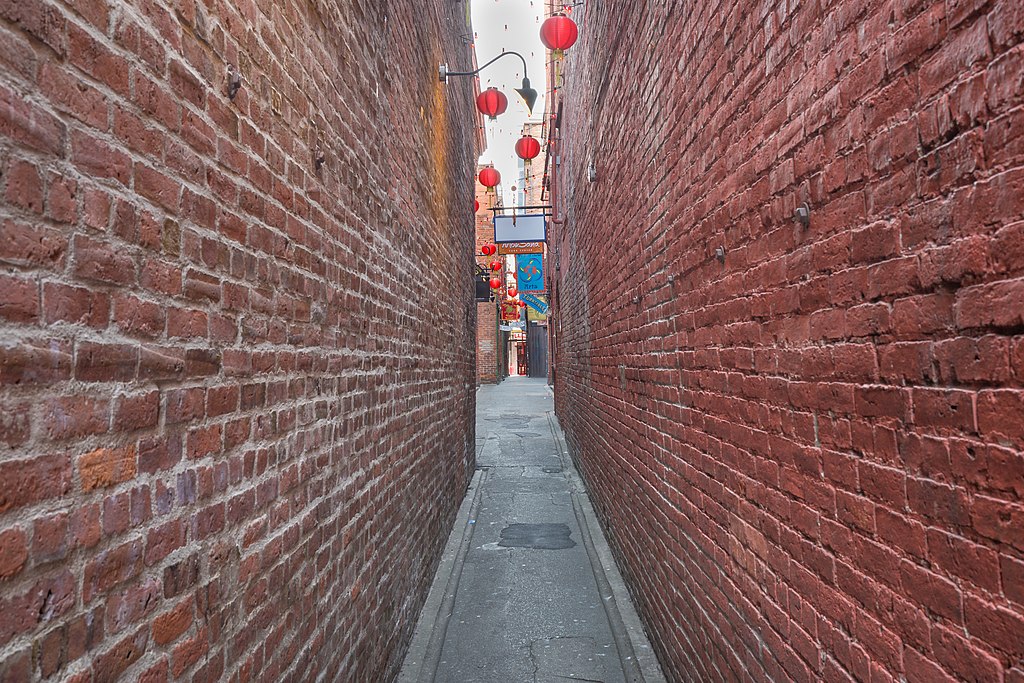 Brick walls frame the sides of a narrow alley. In the distance red Chinese style lanterns and hanging signs for businesses are visible.