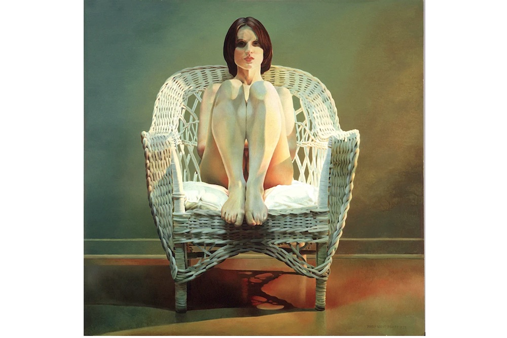 A naked young woman with short dark hair sits on a white cushion in a white wicker chair. She has her knees drawn to her chest and she is looking directly at the viewer.