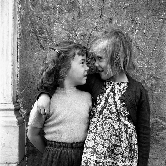 A black and white photograph of two young girls. The blonde girl has her arm around the brunette, there's a look of mischief in her eyes. 