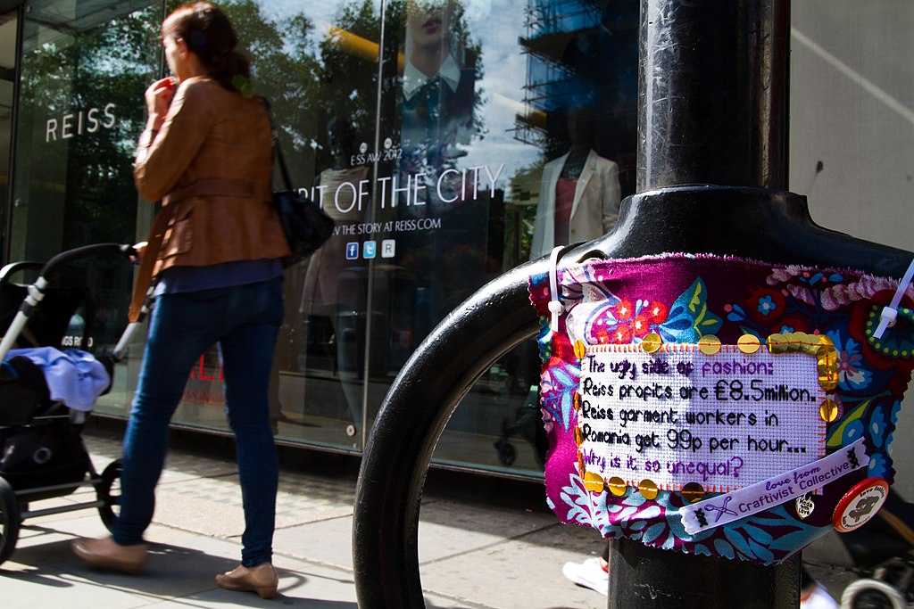 A small hand embroidered sign is attached to a lamp post. It reads:
The ugly side of fashion: Reiss profits are 8.5 million pounds. Reiss garment workers in Romania get 99p per hour... Why is it so unequal?