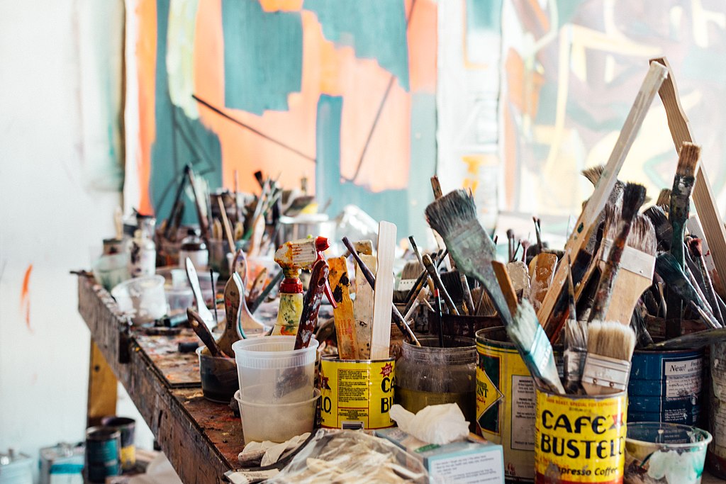 A collection of paint brushes, rags, and other artist's supplies are cluttered on top of a paint-covered workbench.