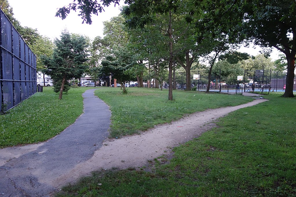 A paved pathway curves to the left between two outdoor sports courts. A desire path connects the court to the paved path. 