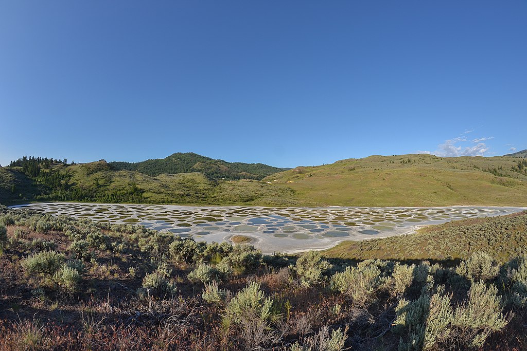 A lake is nestled among brown hills. The surface of the lake has an irregular polkadot pattern with coloured circles amidst a whitish background.