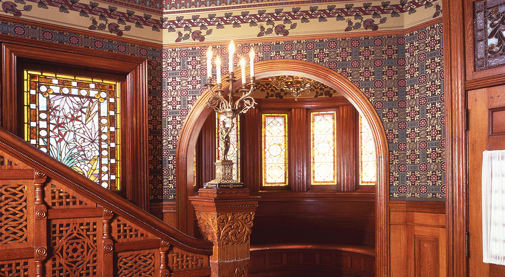 An interior of a Victorian house, showing bold, patterned wallpaper in blue and red, with a floral border and a floral patterned stained glass window. 
