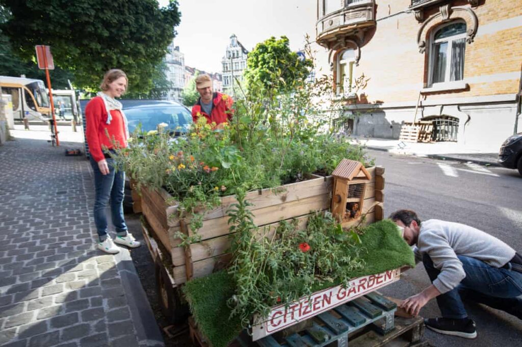 Three people examine a pocket park in a parking space on an urban street. The wooden garden beds are overflowing the lush vegation. The sign reads: Citizen Garden.
